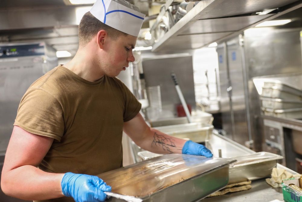 A young male food service worker wearing a white paper chef's hat and olive/tan t-shirt is working in a commercial or military galley kitchen. He is wearing blue nitrile gloves and handling a large stainless steel food service tray, appearing to cover or uncover it with plastic wrap. A tree-branch style tattoo is visible on his right forearm.