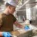 A young male food service worker wearing a white paper chef's hat and olive/tan t-shirt is working in a commercial or military galley kitchen. He is wearing blue nitrile gloves and handling a large stainless steel food service tray, appearing to cover or uncover it with plastic wrap. A tree-branch style tattoo is visible on his right forearm.