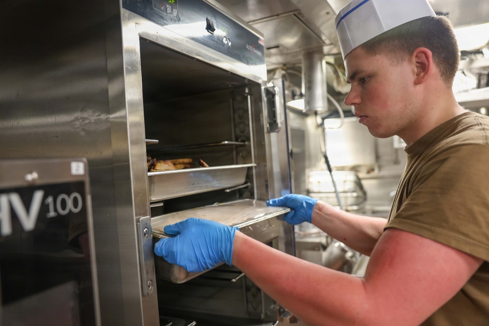 A young male cook wearing a white chef's hat and blue latex gloves is removing or inserting a metal baking tray into a commercial stainless steel oven labeled 'HV100'. He is dressed in a khaki/olive military-style undershirt, suggesting this may be a military galley or mess hall kitchen. The image is taken in a tight, industrial kitchen environment with stainless steel equipment visible in the background.