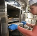 A young male cook wearing a white chef's hat and blue latex gloves is removing or inserting a metal baking tray into a commercial stainless steel oven labeled 'HV100'. He is dressed in a khaki/olive military-style undershirt, suggesting this may be a military galley or mess hall kitchen. The image is taken in a tight, industrial kitchen environment with stainless steel equipment visible in the background.