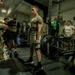 A group of U.S. Army soldiers are conducting a strength training session in a military gym facility. A soldier in the center is performing or preparing for a deadlift using a Hammer Strength barbell loaded with weight plates, while a uniformed soldier with an 82nd Airborne patch appears to be recording or evaluating the performance. The setting appears to be an official physical fitness assessment or training event.