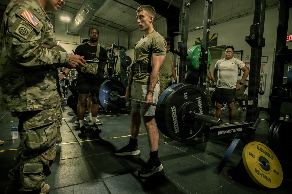 A military fitness assessment is taking place in a gym facility, where a soldier in an olive/tan t-shirt and shorts is performing a deadlift with a Hammer Strength barbell. A uniformed soldier in MultiCam/OCP camouflage with an 82nd Airborne patch and US flag is observing and recording on a clipboard. Several other personnel are visible in the background, suggesting a structured physical fitness test or competition.