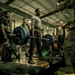A group of military personnel are gathered in a gym facility watching one individual perform a deadlift with a heavily loaded barbell. The lifter is standing on a Hammer Strength platform, gripping a barbell loaded with 45-pound plates. The setting appears to be a formal or competitive strength assessment event on a military installation.