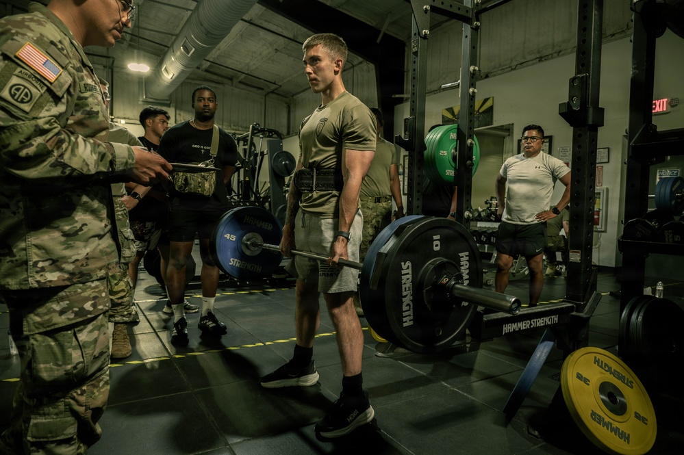 A group of U.S. Army soldiers are conducting a strength training session in a military gym facility. A soldier in the center is performing or preparing for a deadlift using a Hammer Strength barbell loaded with weight plates, while a uniformed soldier with an 82nd Airborne patch appears to be recording or evaluating the performance. The setting appears to be an official physical fitness assessment or training event.