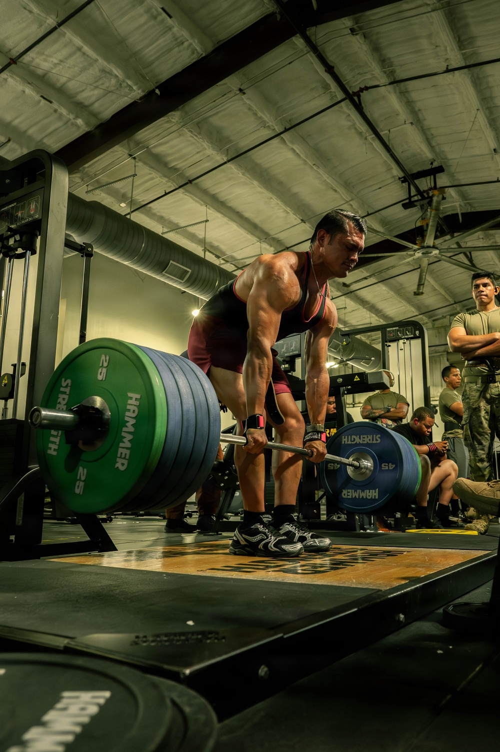 A muscular athlete performs a deadlift in what appears to be a military gym facility, gripping a heavily loaded barbell. The lifter is mid-lift, bent forward with significant weight loaded on the bar, while several observers in military-style clothing watch from the background. The image is taken from a low angle, emphasizing the weight plates and the athlete's muscular physique.