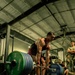 A muscular athlete performs a deadlift in what appears to be a military gym facility, gripping a heavily loaded barbell. The lifter is mid-lift, bent forward with significant weight loaded on the bar, while several observers in military-style clothing watch from the background. The image is taken from a low angle, emphasizing the weight plates and the athlete's muscular physique.