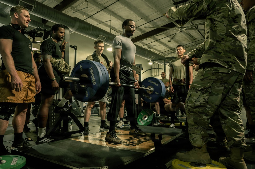 A group of military personnel are gathered in a gym facility watching one individual perform a deadlift with a heavily loaded barbell. The lifter is standing on a Hammer Strength platform, gripping a barbell loaded with 45-pound plates. The setting appears to be a structured fitness assessment or competition event on a military installation.