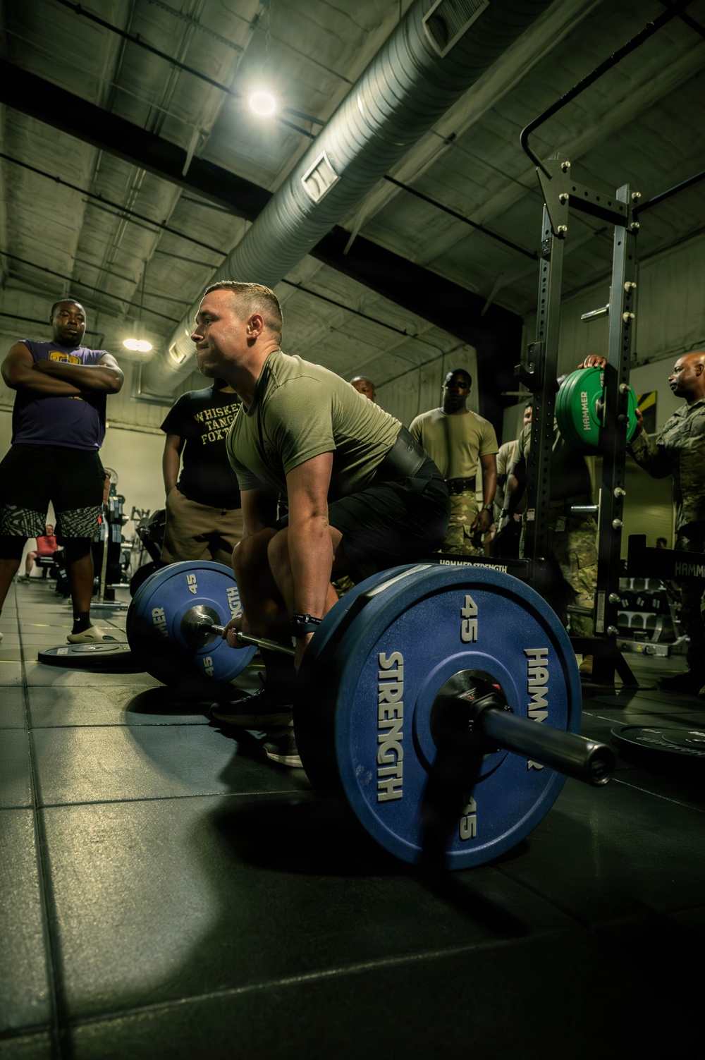 A male athlete in a military-style olive green t-shirt is performing a deadlift with a heavily loaded barbell in an indoor gym facility. The barbell is loaded with Hammer Strength 45-lb bumper plates on both sides. Several onlookers, some in military camouflage uniforms, observe the lift in the background.