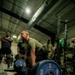 A male athlete in a military-style olive green t-shirt is performing a deadlift with a heavily loaded barbell in an indoor gym facility. The barbell is loaded with Hammer Strength 45-lb bumper plates on both sides. Several onlookers, some in military camouflage uniforms, observe the lift in the background.