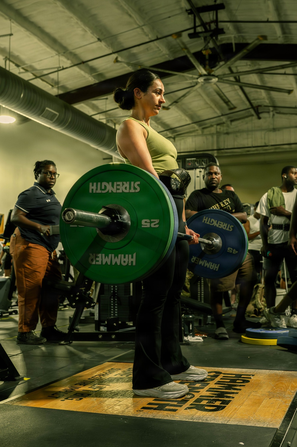A woman in a green tank top and black pants performs a deadlift in a gym setting, holding a loaded barbell with Hammer Strength bumper plates. She is standing on a Hammer Strength branded platform with spectators watching in the background, suggesting a competitive or demonstration lifting event. The barbell is loaded with a 45 lb plate on one side and a 25 lb plate on the other, and she is wearing a lifting belt.
