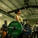 A woman in a green tank top and black pants performs a deadlift in a gym setting, holding a loaded barbell with Hammer Strength bumper plates. She is standing on a Hammer Strength branded platform with spectators watching in the background, suggesting a competitive or demonstration lifting event. The barbell is loaded with a 45 lb plate on one side and a 25 lb plate on the other, and she is wearing a lifting belt.