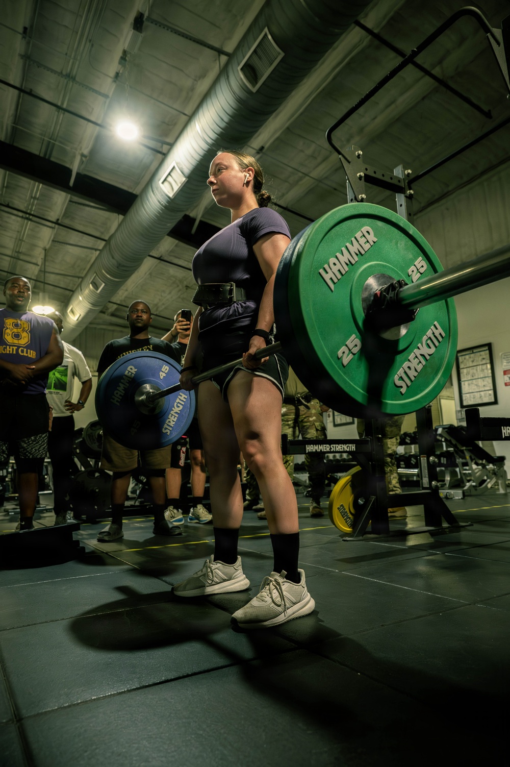 A female athlete is performing a deadlift in an indoor gym facility, holding a loaded barbell with Hammer Strength weight plates. She is wearing a weightlifting belt, dark athletic clothing, black knee-high socks, and white sneakers. A small crowd of spectators is visible in the background, suggesting a competitive or organized lifting event.