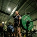 A female athlete is performing a deadlift in an indoor gym facility, holding a loaded barbell with Hammer Strength weight plates. She is wearing a weightlifting belt, dark athletic clothing, black knee-high socks, and white sneakers. A small crowd of spectators is visible in the background, suggesting a competitive or organized lifting event.