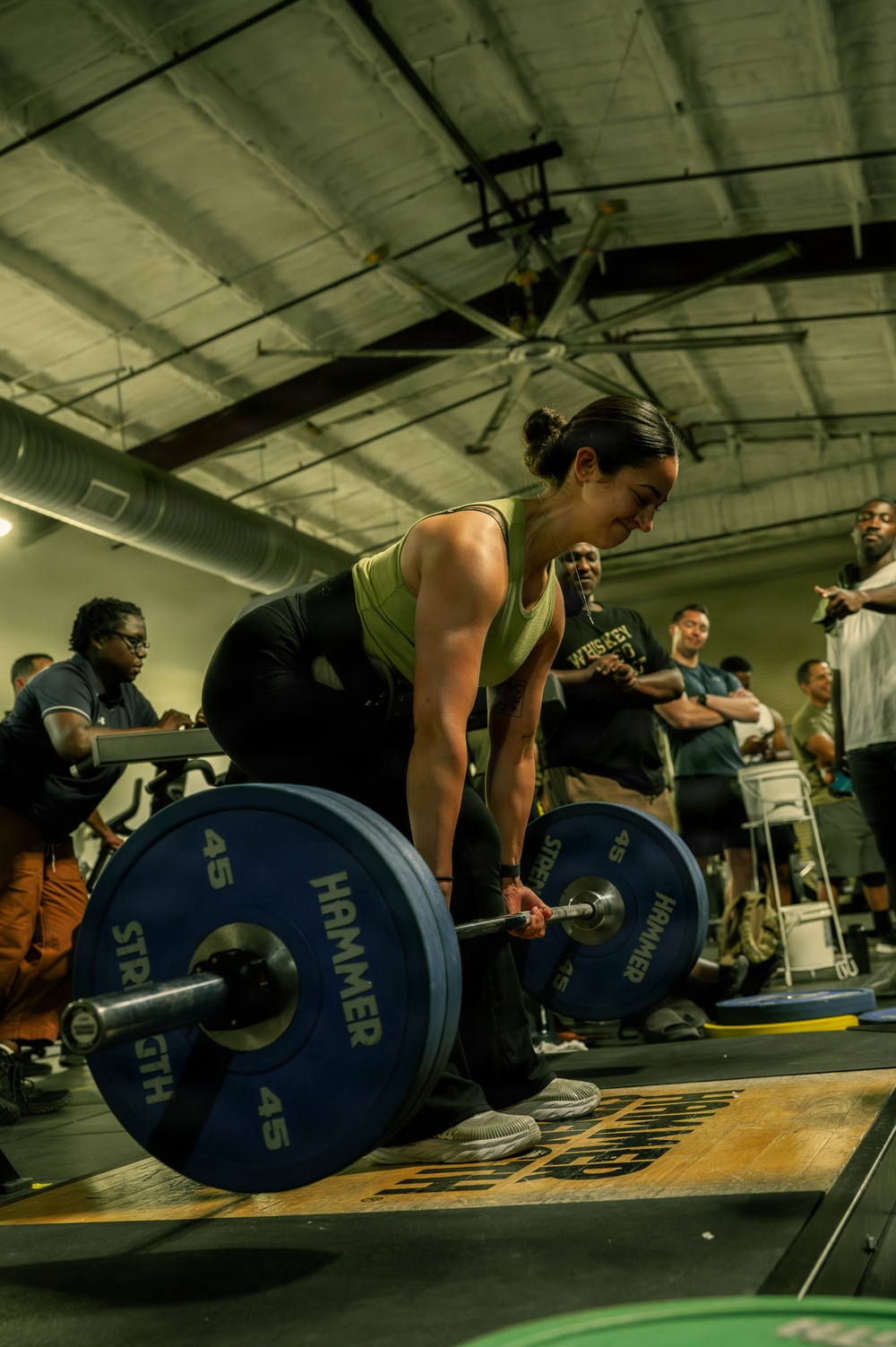 A woman is performing a deadlift in what appears to be a gym competition or event, gripping a loaded barbell with 45-pound Hammer Strength bumper plates on each side. She is wearing a green tank top and black leggings, positioned in the starting deadlift stance with spectators watching in the background. The setting is an industrial-style gym with exposed ductwork and large ceiling fans.