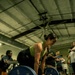 A woman is performing a deadlift in what appears to be a gym competition or event, gripping a loaded barbell with 45-pound Hammer Strength bumper plates on each side. She is wearing a green tank top and black leggings, positioned in the starting deadlift stance with spectators watching in the background. The setting is an industrial-style gym with exposed ductwork and large ceiling fans.
