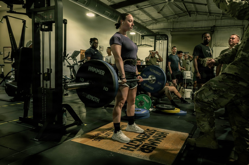 A woman is performing a deadlift in a military gym facility, standing on a Hammer Strength branded platform while holding a loaded barbell. She is surrounded by a group of onlookers, several of whom appear to be in military attire including camouflage uniforms. The setting appears to be an organized lifting event or competition within a military installation.