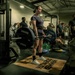 A woman is performing a deadlift in a military gym facility, standing on a Hammer Strength branded platform while holding a loaded barbell. She is surrounded by a group of onlookers, several of whom appear to be in military attire including camouflage uniforms. The setting appears to be an organized lifting event or competition within a military installation.