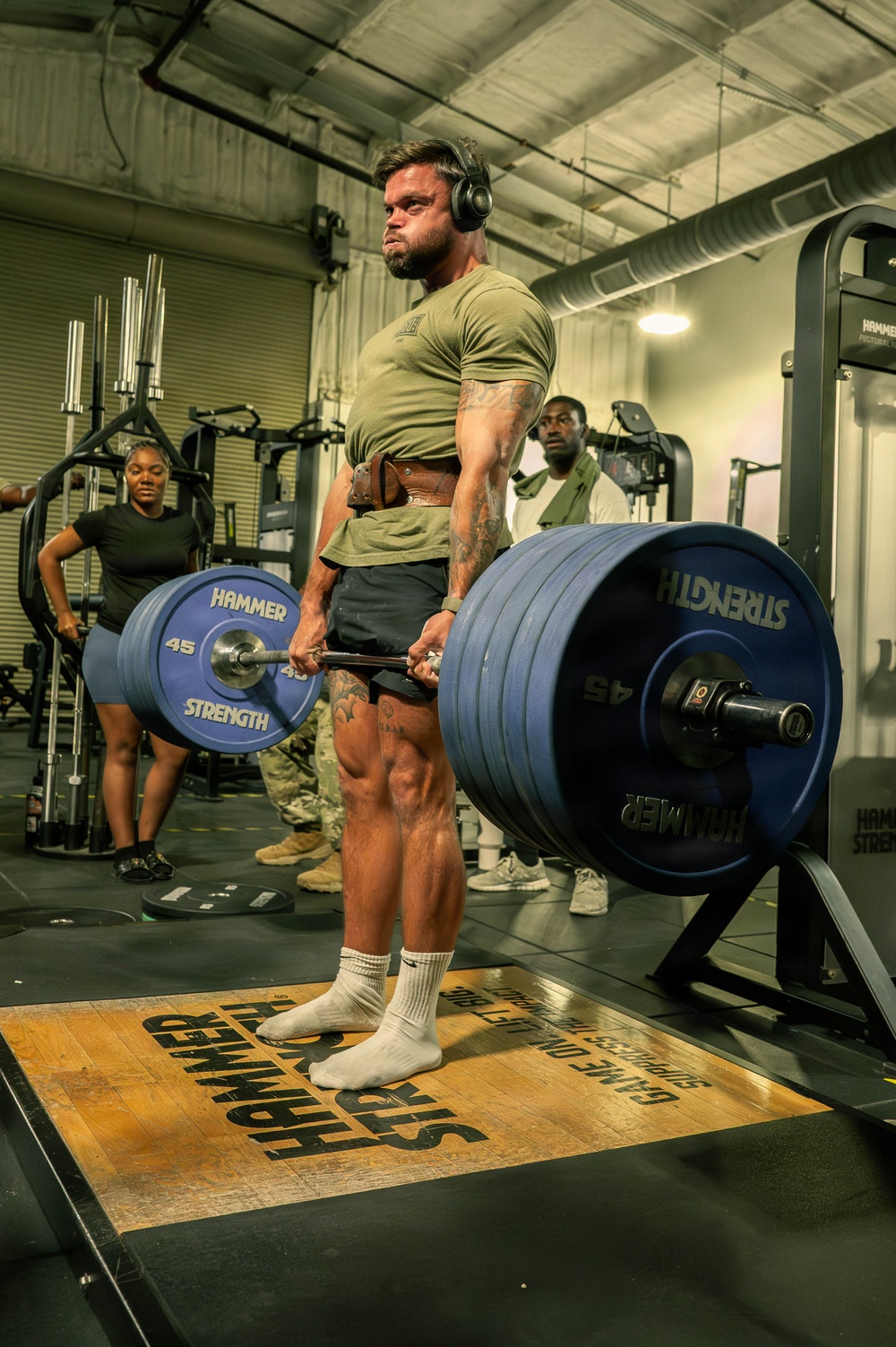 A heavily muscular man wearing a green military-style t-shirt, shorts, white socks, and over-ear headphones is performing a deadlift with a heavily loaded barbell in an indoor gym. The barbell is loaded with multiple Hammer Strength 45-lb bumper plates on each side. Several other individuals in military-style attire are visible in the background observing or working out.