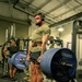 A heavily muscular man wearing a green military-style t-shirt, shorts, white socks, and over-ear headphones is performing a deadlift with a heavily loaded barbell in an indoor gym. The barbell is loaded with multiple Hammer Strength 45-lb bumper plates on each side. Several other individuals in military-style attire are visible in the background observing or working out.