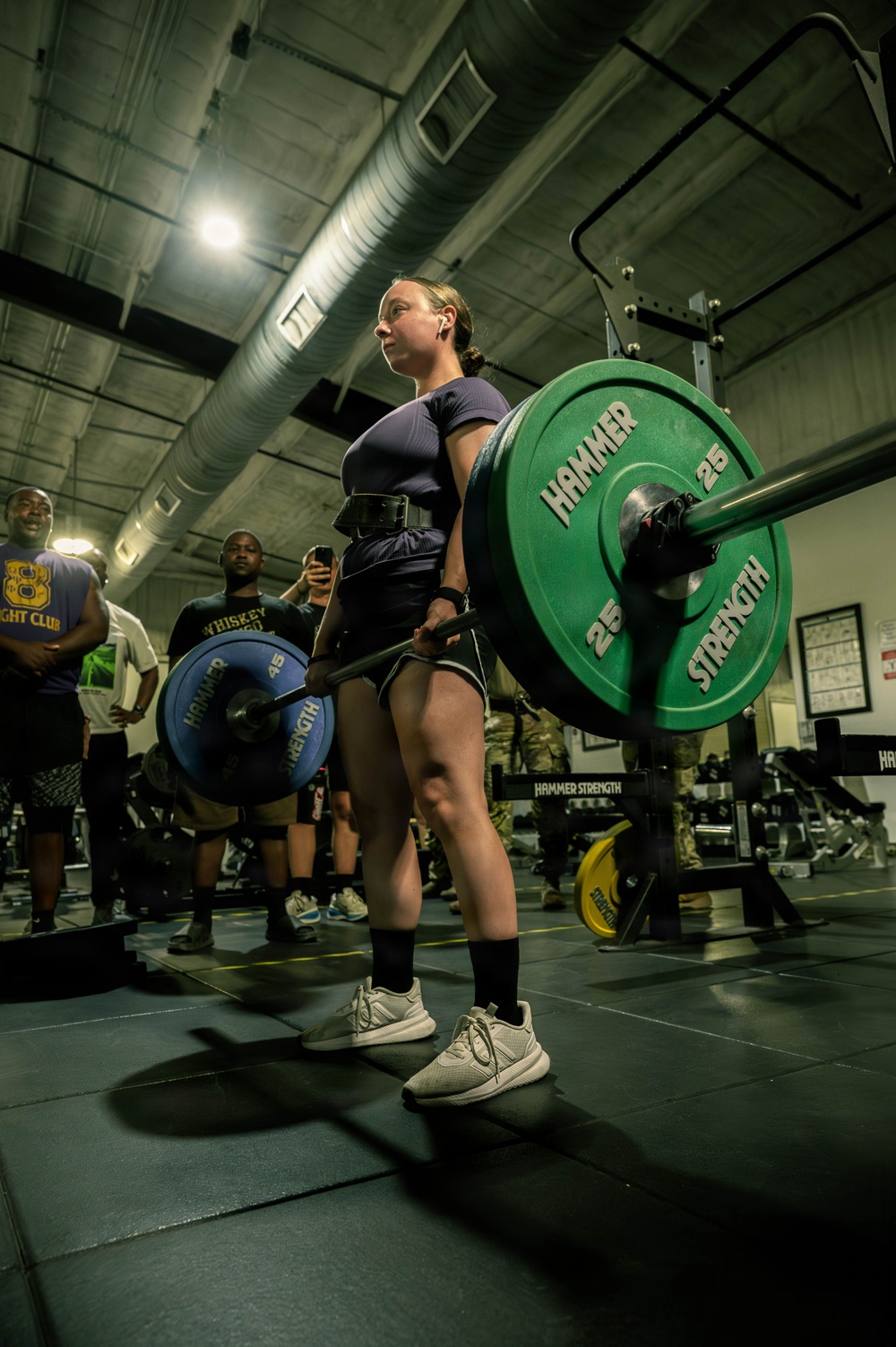 A woman is performing a deadlift in an indoor gym facility, holding a loaded barbell with Hammer Strength weight plates. She is wearing a weightlifting belt, dark shorts, a navy t-shirt, and white sneakers, with several spectators watching in the background. The setting appears to be a military or institutional gym based on the equipment branding and environment.