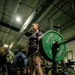 A woman is performing a deadlift in an indoor gym facility, holding a loaded barbell with Hammer Strength weight plates. She is wearing a weightlifting belt, dark shorts, a navy t-shirt, and white sneakers, with several spectators watching in the background. The setting appears to be a military or institutional gym based on the equipment branding and environment.