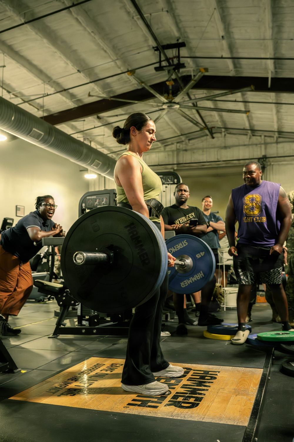 A woman is performing a deadlift at what appears to be 'Hammer Strength' gym, lifting a heavily loaded barbell with multiple 45-pound plates. Several spectators in the background are visibly cheering and encouraging her, suggesting this may be a competition or a notable personal record attempt. The gym has an industrial aesthetic with exposed ductwork and ceiling fans.