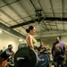A woman is performing a deadlift at what appears to be 'Hammer Strength' gym, lifting a heavily loaded barbell with multiple 45-pound plates. Several spectators in the background are visibly cheering and encouraging her, suggesting this may be a competition or a notable personal record attempt. The gym has an industrial aesthetic with exposed ductwork and ceiling fans.