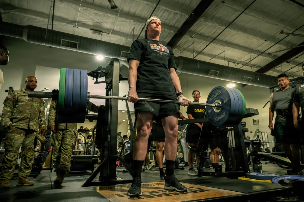 A person in a black t-shirt and shorts is performing a deadlift with a heavily loaded barbell in a military gym facility. Several onlookers in U.S. Army combat uniforms (ACUs) and PT gear observe the lift. The setting appears to be an official Army fitness event or competition based on the branded equipment and uniformed personnel present.