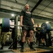 A person in a black t-shirt and shorts is performing a deadlift with a heavily loaded barbell in a military gym facility. Several onlookers in U.S. Army combat uniforms (ACUs) and PT gear observe the lift. The setting appears to be an official Army fitness event or competition based on the branded equipment and uniformed personnel present.