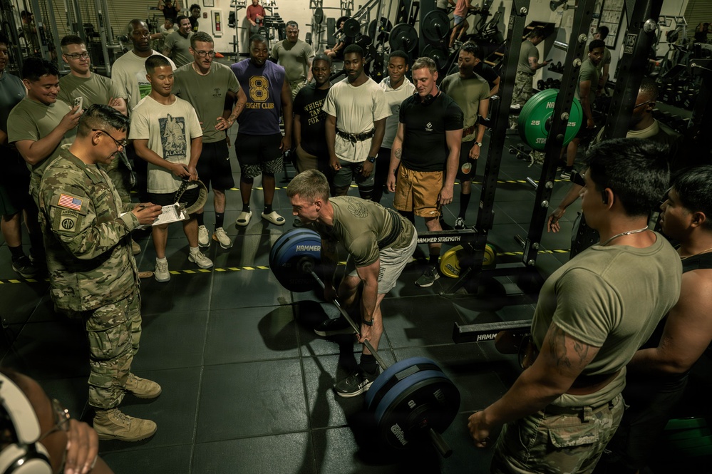 A military fitness event is taking place inside a gym, where a soldier is performing a deadlift with Hammer Strength weight plates while surrounded by a large crowd of observers. A uniformed soldier in camouflage (ACU/OCP pattern) stands to the left taking notes on a clipboard, suggesting this is a formal assessment or competition. The setting appears to be a well-equipped military gymnasium with power racks and weight equipment visible in the background.
