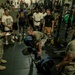 A military fitness event is taking place inside a gym, where a soldier is performing a deadlift with Hammer Strength weight plates while surrounded by a large crowd of observers. A uniformed soldier in camouflage (ACU/OCP pattern) stands to the left taking notes on a clipboard, suggesting this is a formal assessment or competition. The setting appears to be a well-equipped military gymnasium with power racks and weight equipment visible in the background.