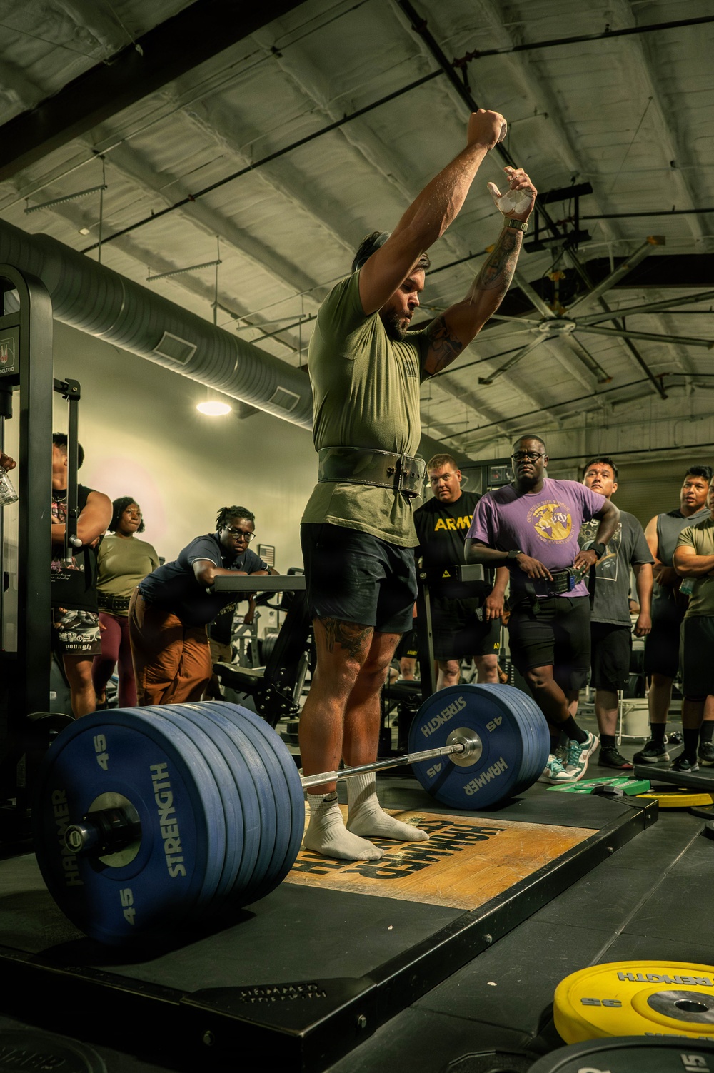 A powerlifter has just completed a deadlift at what appears to be a competitive lifting event or gym meet, raising his hands in a celebratory gesture. He is standing on a marked lifting platform surrounded by spectators and other athletes. The setting is an industrial-style gym with exposed ductwork and ceiling fans.