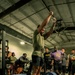 A powerlifter has just completed a deadlift at what appears to be a competitive lifting event or gym meet, raising his hands in a celebratory gesture. He is standing on a marked lifting platform surrounded by spectators and other athletes. The setting is an industrial-style gym with exposed ductwork and ceiling fans.