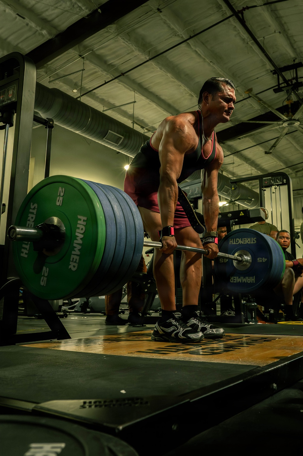 A heavily muscular individual is performing a deadlift in a commercial gym, gripping a heavily loaded barbell with visible strain on their face. The barbell is loaded with Hammer Strength plates, including 45 lb and 25 lb plates on each side. The low-angle shot emphasizes the athlete's musculature and the weight being lifted, with other gym-goers visible in the background.
