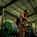 A heavily muscular individual is performing a deadlift in a commercial gym, gripping a heavily loaded barbell with visible strain on their face. The barbell is loaded with Hammer Strength plates, including 45 lb and 25 lb plates on each side. The low-angle shot emphasizes the athlete's musculature and the weight being lifted, with other gym-goers visible in the background.