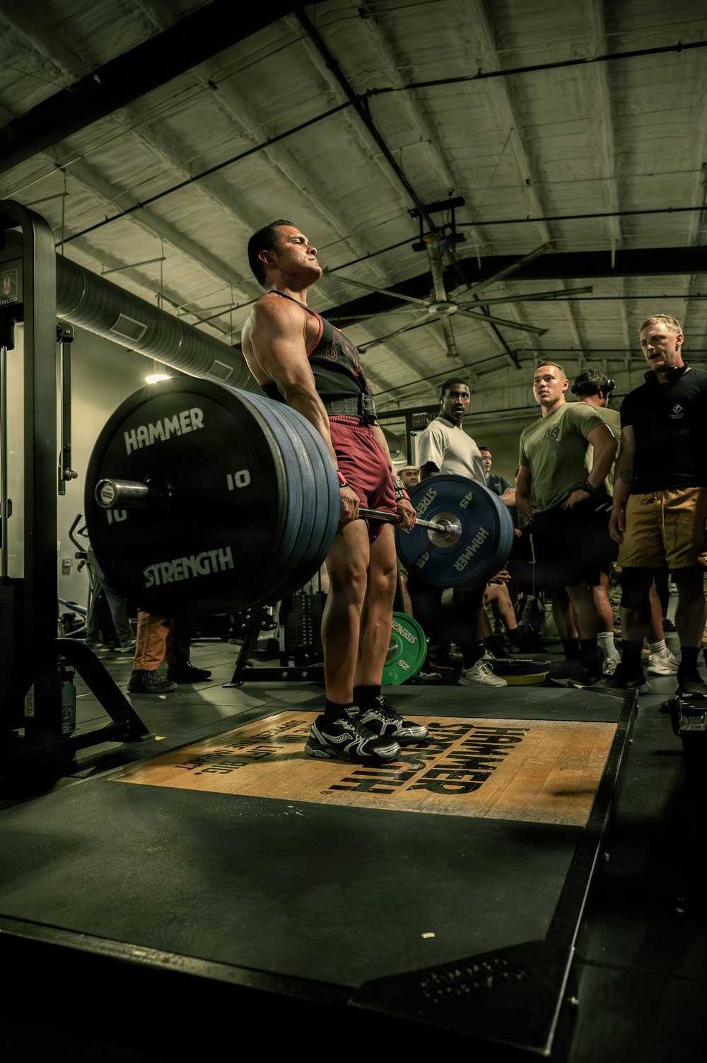 A muscular athlete performs a deadlift on a marked platform in an indoor gym facility, surrounded by a group of spectators. The lifter is using a heavily loaded barbell with Hammer Strength bumper plates, wearing a lifting belt and red shorts. The setting appears to be a competitive or demonstration lifting event with multiple observers watching intently.