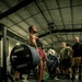 A muscular athlete performs a deadlift on a marked platform in an indoor gym facility, surrounded by a group of spectators. The lifter is using a heavily loaded barbell with Hammer Strength bumper plates, wearing a lifting belt and red shorts. The setting appears to be a competitive or demonstration lifting event with multiple observers watching intently.
