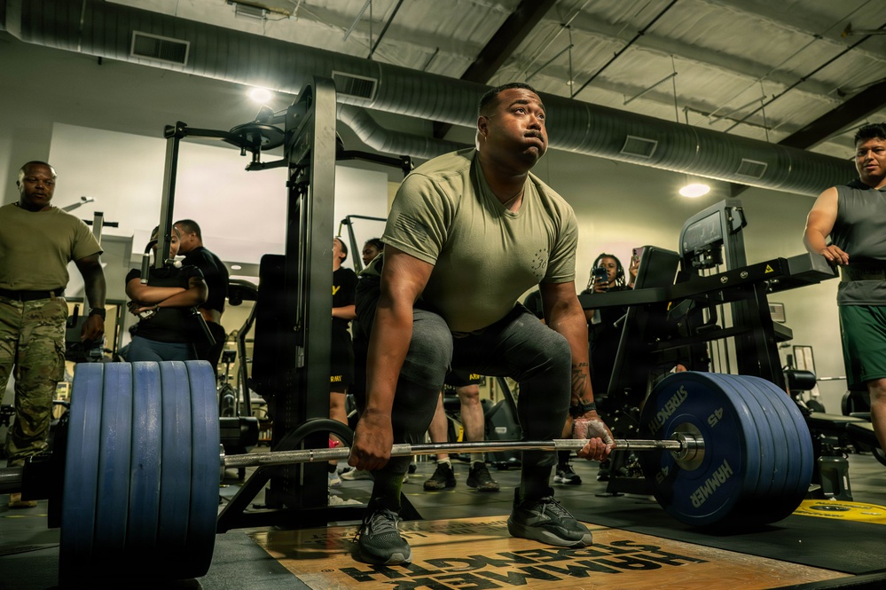 A muscular individual in an olive/tan military-style t-shirt is performing a deadlift with a heavily loaded barbell in a gym setting. Multiple observers, several wearing military uniforms and Army-branded clothing, are watching from the background. The setting appears to be a military fitness facility based on the 'ARMY' text visible on equipment and attire.