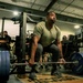 A muscular individual in an olive/tan military-style t-shirt is performing a deadlift with a heavily loaded barbell in a gym setting. Multiple observers, several wearing military uniforms and Army-branded clothing, are watching from the background. The setting appears to be a military fitness facility based on the 'ARMY' text visible on equipment and attire.