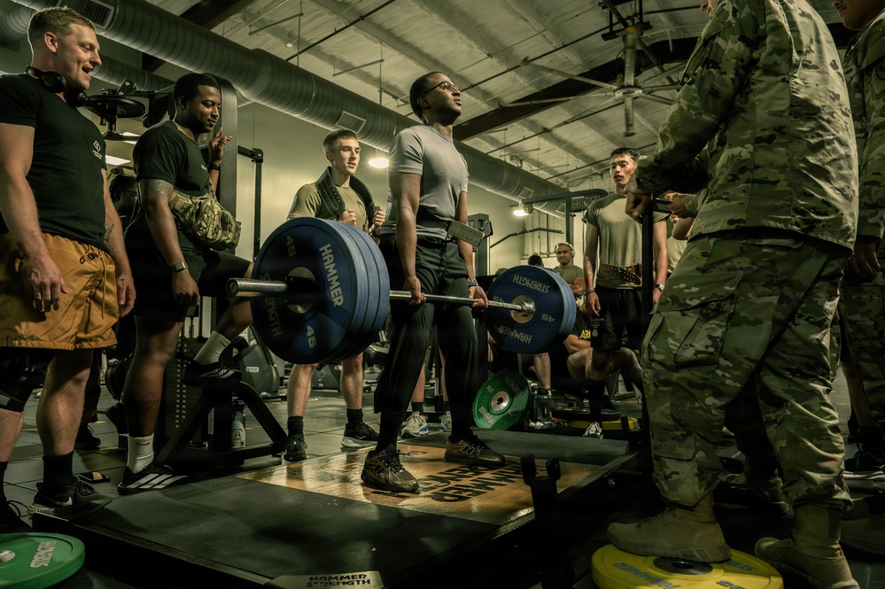 A group of military personnel are gathered in a gym facility, observing a soldier performing a deadlift with a heavily loaded barbell. The lifter is standing on a lifting platform surrounded by onlookers in various military and workout attire. The setting appears to be a military fitness center, possibly during a formal strength assessment or competition.