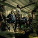 A group of military personnel are gathered in a gym facility, observing a soldier performing a deadlift with a heavily loaded barbell. The lifter is standing on a lifting platform surrounded by onlookers in various military and workout attire. The setting appears to be a military fitness center, possibly during a formal strength assessment or competition.