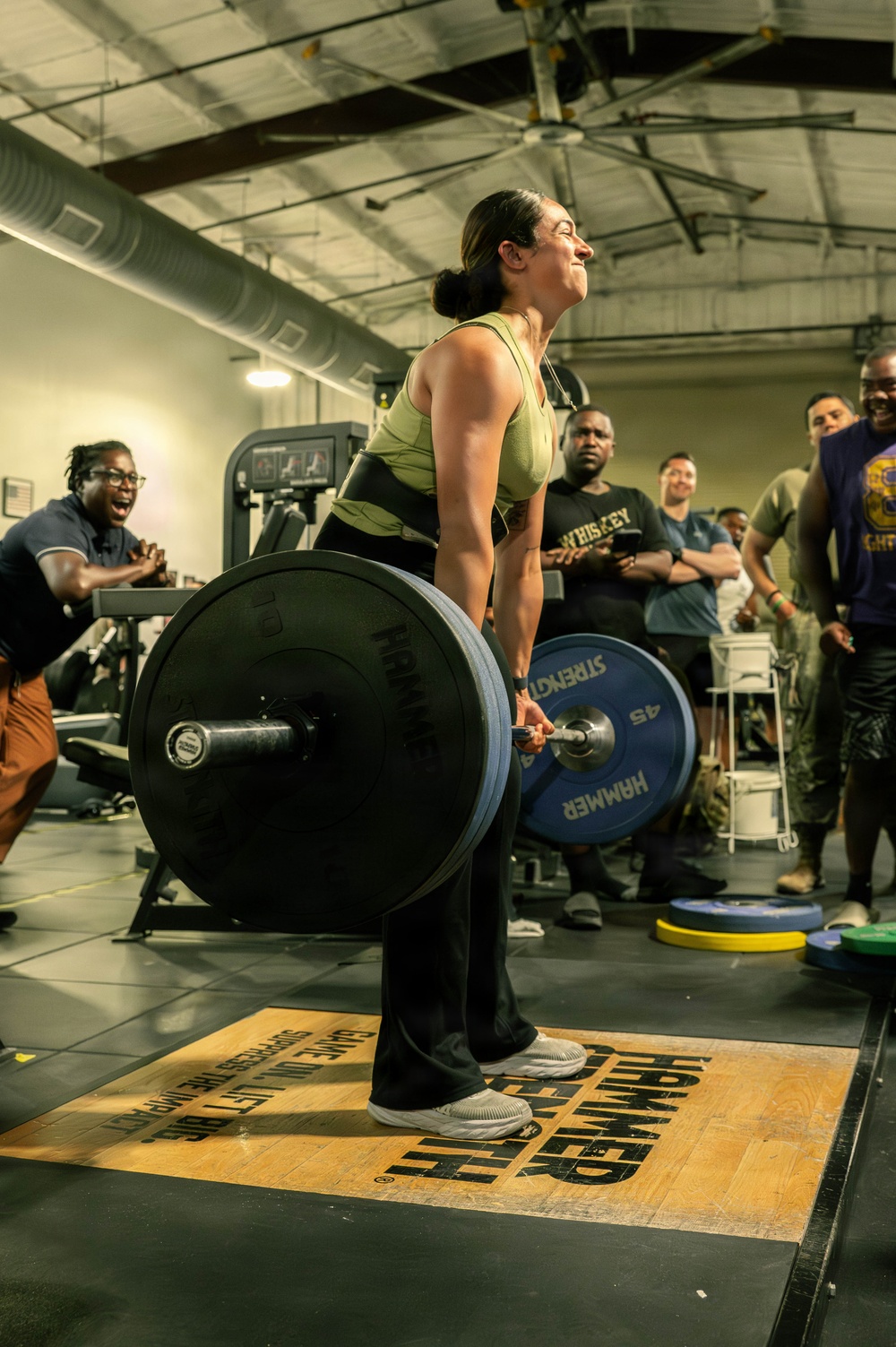 A woman is performing a deadlift in what appears to be a gym competition or event, successfully lifting a heavily loaded barbell. She is wearing a green sports bra and black pants, standing on a Hammer Strength branded lifting platform. A crowd of spectators surrounds her, visibly cheering and reacting with excitement.