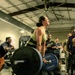 A woman is performing a deadlift in what appears to be a gym competition or event, successfully lifting a heavily loaded barbell. She is wearing a green sports bra and black pants, standing on a Hammer Strength branded lifting platform. A crowd of spectators surrounds her, visibly cheering and reacting with excitement.