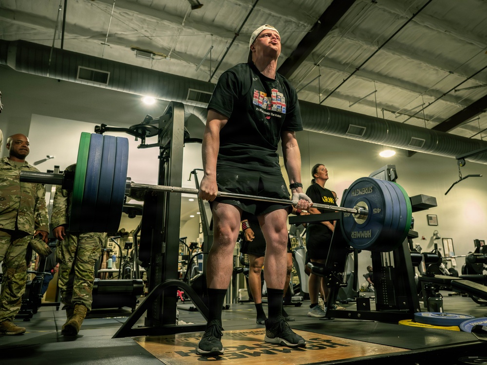 A person in a black t-shirt and shorts is performing a deadlift with a heavily loaded barbell in a military gym facility. Several individuals in military camouflage uniforms are observing in the background, suggesting this may be a fitness event or competition. The lifter appears to be exerting maximum effort, with head tilted back and an expression of intense strain.