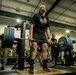 A person in a black t-shirt and shorts is performing a deadlift with a heavily loaded barbell in a military gym facility. Several individuals in military camouflage uniforms are observing in the background, suggesting this may be a fitness event or competition. The lifter appears to be exerting maximum effort, with head tilted back and an expression of intense strain.