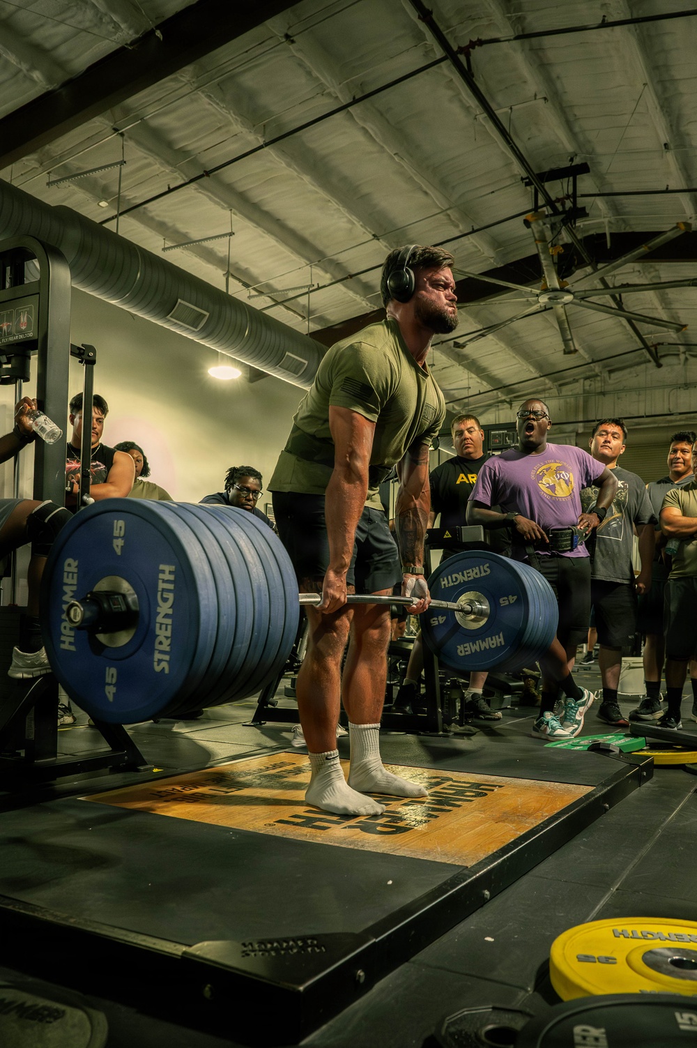 A muscular man wearing a green t-shirt, shorts, white socks, and over-ear headphones is performing a deadlift on a Hammer Strength platform in a gym setting. The barbell is loaded with multiple 45 lb Hammer Strength bumper plates on each side. A crowd of spectators stands in the background watching the lift, suggesting a competitive or demonstration event.
