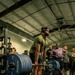 A muscular man wearing a green t-shirt, shorts, white socks, and over-ear headphones is performing a deadlift on a Hammer Strength platform in a gym setting. The barbell is loaded with multiple 45 lb Hammer Strength bumper plates on each side. A crowd of spectators stands in the background watching the lift, suggesting a competitive or demonstration event.