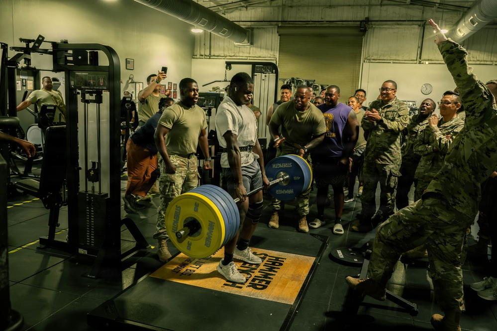 A military gym setting where a soldier in a white t-shirt is performing a heavy deadlift on a Hammer Strength platform, surrounded by a large crowd of fellow service members in military uniforms who are cheering and reacting enthusiastically. The lifter appears to be mid-lift with a heavily loaded barbell featuring yellow and blue weight plates. The atmosphere is energetic and celebratory, suggesting a fitness competition or challenge event.