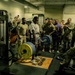 A military gym setting where a soldier in a white t-shirt is performing a heavy deadlift on a Hammer Strength platform, surrounded by a large crowd of fellow service members in military uniforms who are cheering and reacting enthusiastically. The lifter appears to be mid-lift with a heavily loaded barbell featuring yellow and blue weight plates. The atmosphere is energetic and celebratory, suggesting a fitness competition or challenge event.