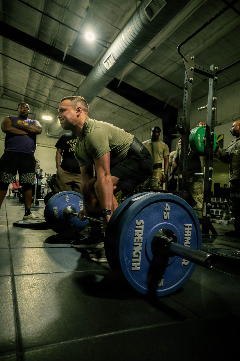 A person in a military-style olive green t-shirt is performing a deadlift with a heavily loaded barbell in an indoor gym facility. The barbell is loaded with Hammer Strength 45-lb blue bumper plates on both sides. Several onlookers, some in military camouflage uniforms, observe the lift in the background.